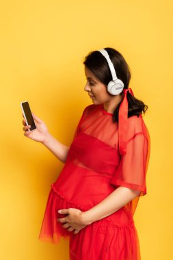 Kablosuz kulaklıklı genç bir kadın akıllı telefon tutuyor ve sarıda karnına dokunuyor.