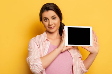 Kumral kadın elinde tablet tutarken boş ekran ile sarı kameraya bakıyor.