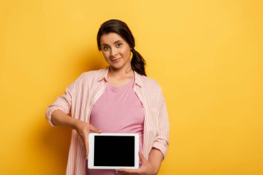Genç hamile bir kadın, sarı ekranda kameraya bakarken boş ekranla dijital tablet gösteriyor.