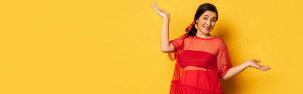panoramic shot of pregnant woman in red tunic standing with open arms while looking at camera on yellow