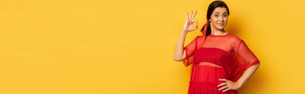 horizontal image of pregnant woman in red tunic showing okay gesture while holding hand on hip on yellow