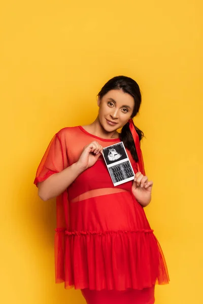 黄色のカメラを見ながら超音波スキャンを示す赤いチュニックの若い妊婦 — ストック写真