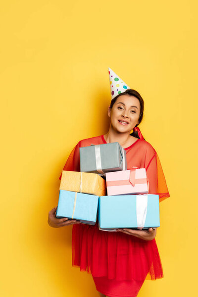 young pregnant woman in party cap holding stack of gift boxes on yellow