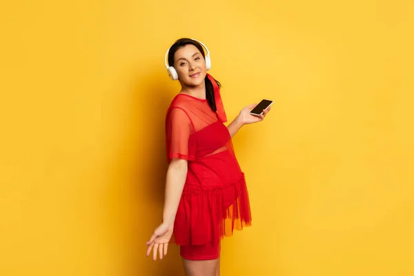 Ciężarna Kobieta Słuchawkach Bezprzewodowych Trzymając Smartfona Stojąc Żółtym — Zdjęcie stockowe