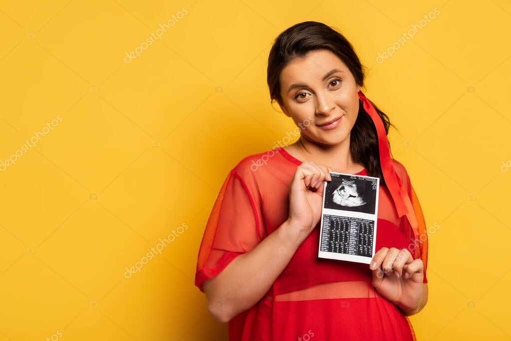 Young pregnant woman in red outfit holding ultrasound scan while looking at camera on yellow