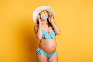Şapkalı, güneş gözlüklü ve sarı maskeli hamile bir kadın.