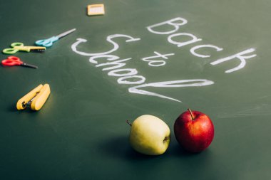 selective focus of delicious apples near back to school inscription and school supplies on green chalkboard clipart