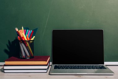 boş ekranlı dizüstü bilgisayar ve yeşil karatahtanın yanındaki kitap yığınında kırtasiye malzemesi olan kalem tutacağı