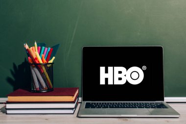 KYIV, UKRAINE - 7 Temmuz 2020: Yeşil karatahtanın yakınındaki ders kitapları yığınında kalem tutacağının yanında HBO web sitesi bulunan dizüstü bilgisayar