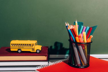 school bus model on staked books near pen holder with stationery on notebook near green chalkboard clipart