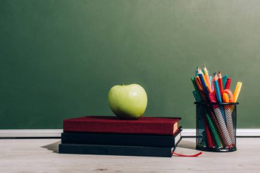 ripe apple on books near pen holder with school supplies on desk near green chalkboard clipart