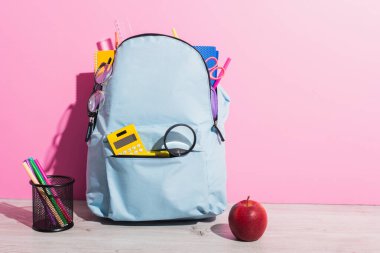Okul çantası dolusu kırtasiye malzemesi. Pembe kalemli, elma ve kalem tutacağının yanında.