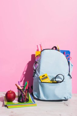 Not defterlerinin yanında okul gereçleri olan mavi bir sırt çantası, pembe kalemli elma ve kalem tutacağı.