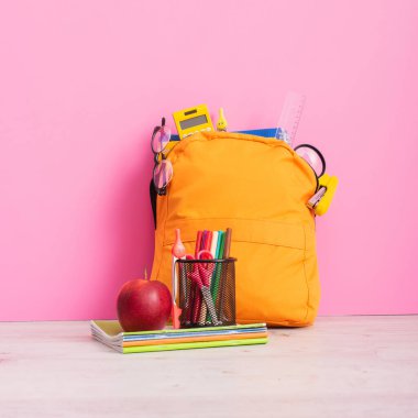 Okul malzemeleriyle dolu sarı sırt çantası defterlerin yanında, keçeli kalemlerle dolu kalem tutacağı, makas ve pembe üzerine olgun elma.