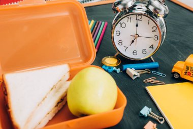 Lezzetli sandviçlerle dolu yemek kutusunun seçici odağı ve antika çalar saatin yanında olgun elma ve siyah karatahtanın üzerinde okul malzemeleri.