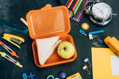 Olgun elmalı beslenme çantasının üst görüntüsü ve antika çalar saatin yanında sandviçler ve siyah karatahtanın üzerinde okul kırtasiyesi.