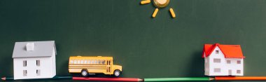 top view of toy school bus and house models on road made of color pencils, and sun made of magnets on green chalkboard, horizontal image clipart