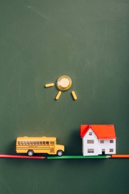 top view of toy school bus on road made of color pencils near house model, and sun made of magnets on green chalkboard clipart