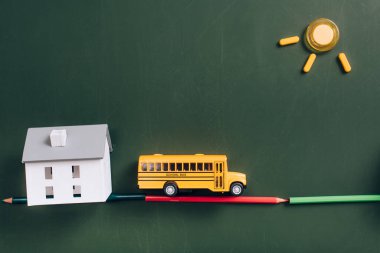 Yol üzerindeki sarı okul otobüsünün üst görüntüsü renkli kalemler, ev modeli ve yeşil karatahtanın üzerindeki mıknatıslardan yapılmış güneş.