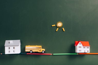 top view of toy school bus on road made of color pencils, house models and sun made of magnets on green chalkboard clipart