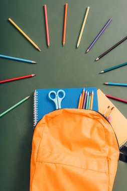 Yeşil karatahtadaki renkli kalemlerin yanında okul gereçleri olan sarı sırt çantasının üst görüntüsü