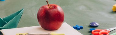 selective focus of delicious apple on book near school stationery on green chalkboard, panoramic concept clipart
