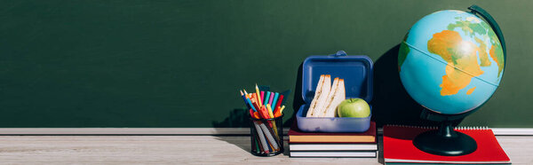 horizontal image of globe near lunch box on books and pen holder near green chalkboard