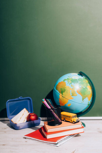 globe near school bus model and pen holder on books near lunch box on desk near green chalkboard