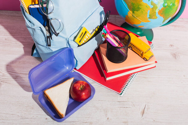 high angle view of blue backpack with school supplies near lunch box, globe, books, school bus model and apple on pink