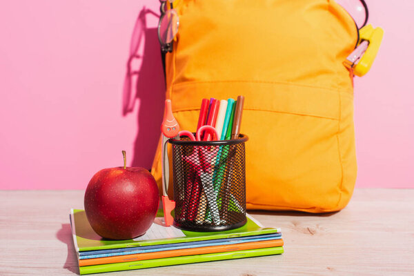 packed school backpack near notebooks, pen holder with felt pens, scissors and ripe apple on pink