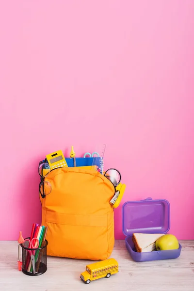 午餐盒附近装有学习用品的黄色背包 校车模型和粉红毛笔笔架 — 图库照片