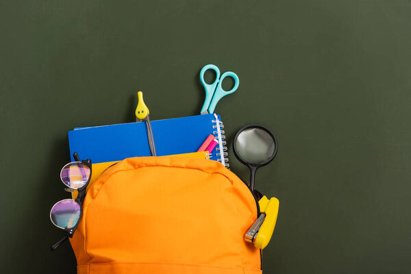 top view of yellow backpack full of school stationery on green chalkboard