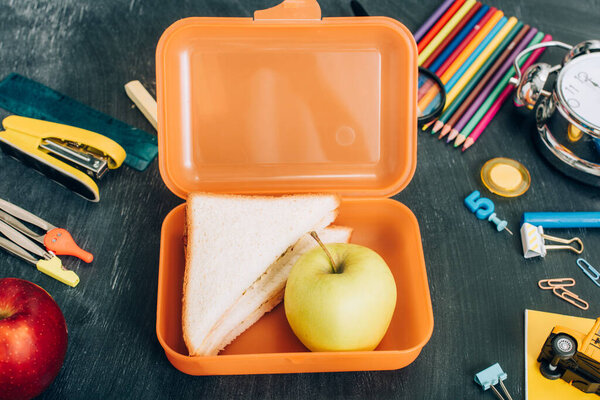 вид на обеденную коробку с бутербродами и спелое яблоко возле школьных канцелярских принадлежностей на черной доске