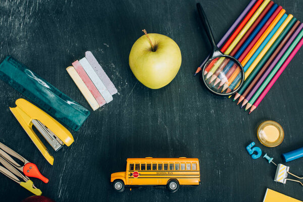 top view of school bus model, whole apple and school stationery on black chalkboard 