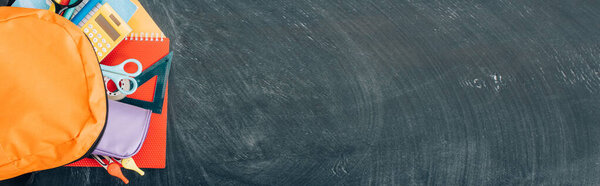 horizontal image of backpack with multicolored school supplies on black chalkboard, top view