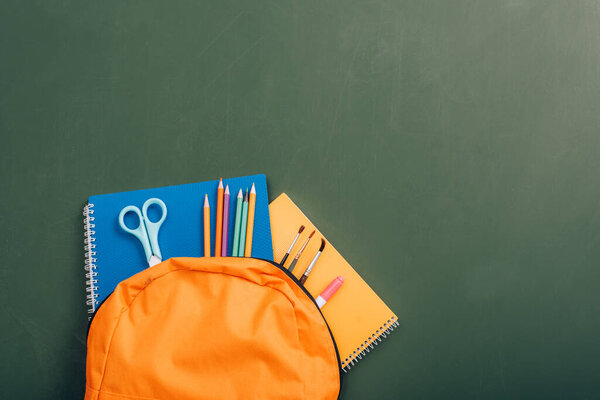 school backpack with colorful copy books, color pencils, scissors and paintbrushes on green chalkboard