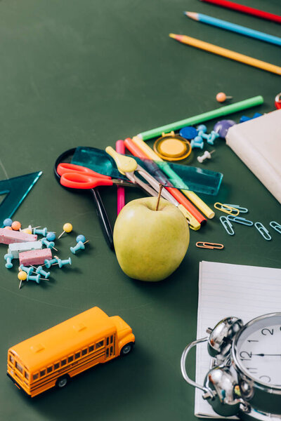 selective focus of ripe apple, school bus model and vintage alarm clock near school supplies on green chalkboard, high angle view