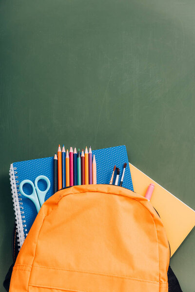 top view of yellow backpack with notebooks, scissors, paintbrushes and color pencils on green chalkboard