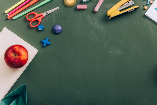top view of tasty apple on book near school stationery on green chalkboard