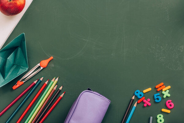 top view of school stationery, paper boat and whole apple on green chalkboard