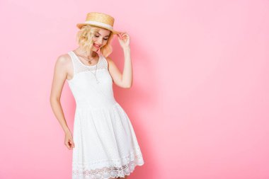 Beyaz elbiseli genç kadın hasır şapkaya dokunuyor ve pembeye bakıyor. 