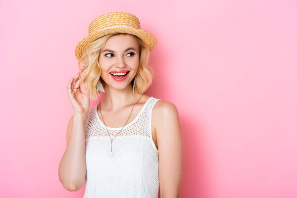 young woman in straw hat touching hair and looking away on pink 