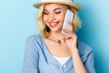 Hasır şapkalı genç kadın mavi telefonda akıllı telefondan konuşuyor. 