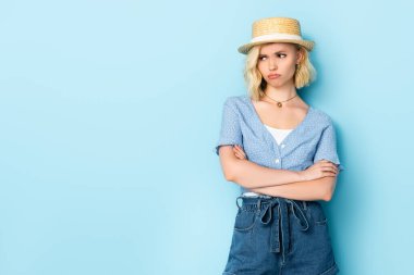 displeased young woman in straw hat standing with crossed arms and looking away on blue  clipart
