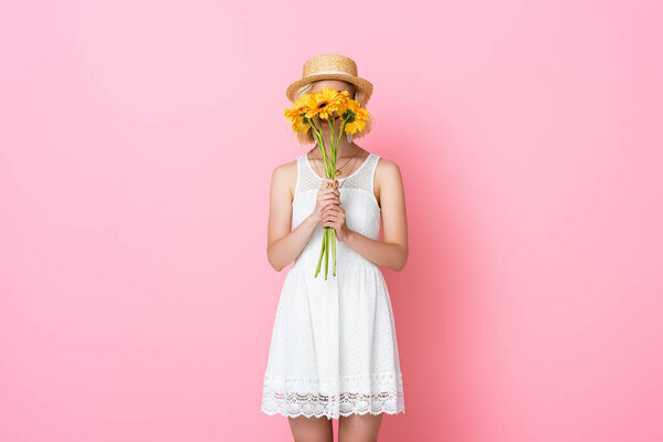 young woman in straw hat and white dress covering face with yellow flowers on pink 