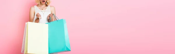 horizontal crop of surprised woman in white dress holding shopping bags on pink