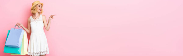 панорамная концепция женщины в соломенной шляпе, указывающей пальцем и держащей сумки с покупками на розовом 