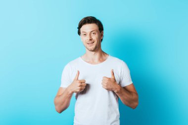 Young man in white t-shirt showing thumbs up on blue background clipart