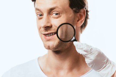 Dermatolojist, beyaz tenli hastanın yanağına yakın büyüteç tutuyor.