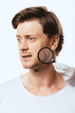 Dermatolojist, beyaz tenli hastanın yanağına yakın büyüteç tutuyor.